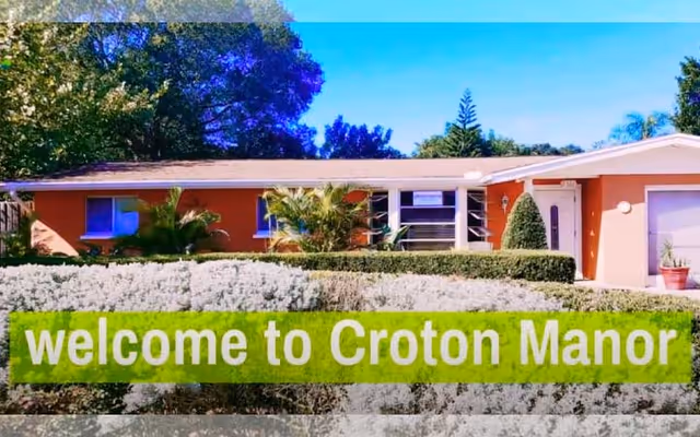 Exterior view of a single-story building with an orange facade, surrounded by green bushes and trees under a clear blue sky. Text overlay at the bottom reads 'welcome to Croton Manor'.