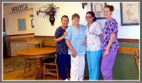 Four women standing together inside a room with wooden chairs and a round table. The women are wearing colorful scrubs and smiling. The room has framed pictures on the wall and a sign that reads 'WERLEY'S LA'.