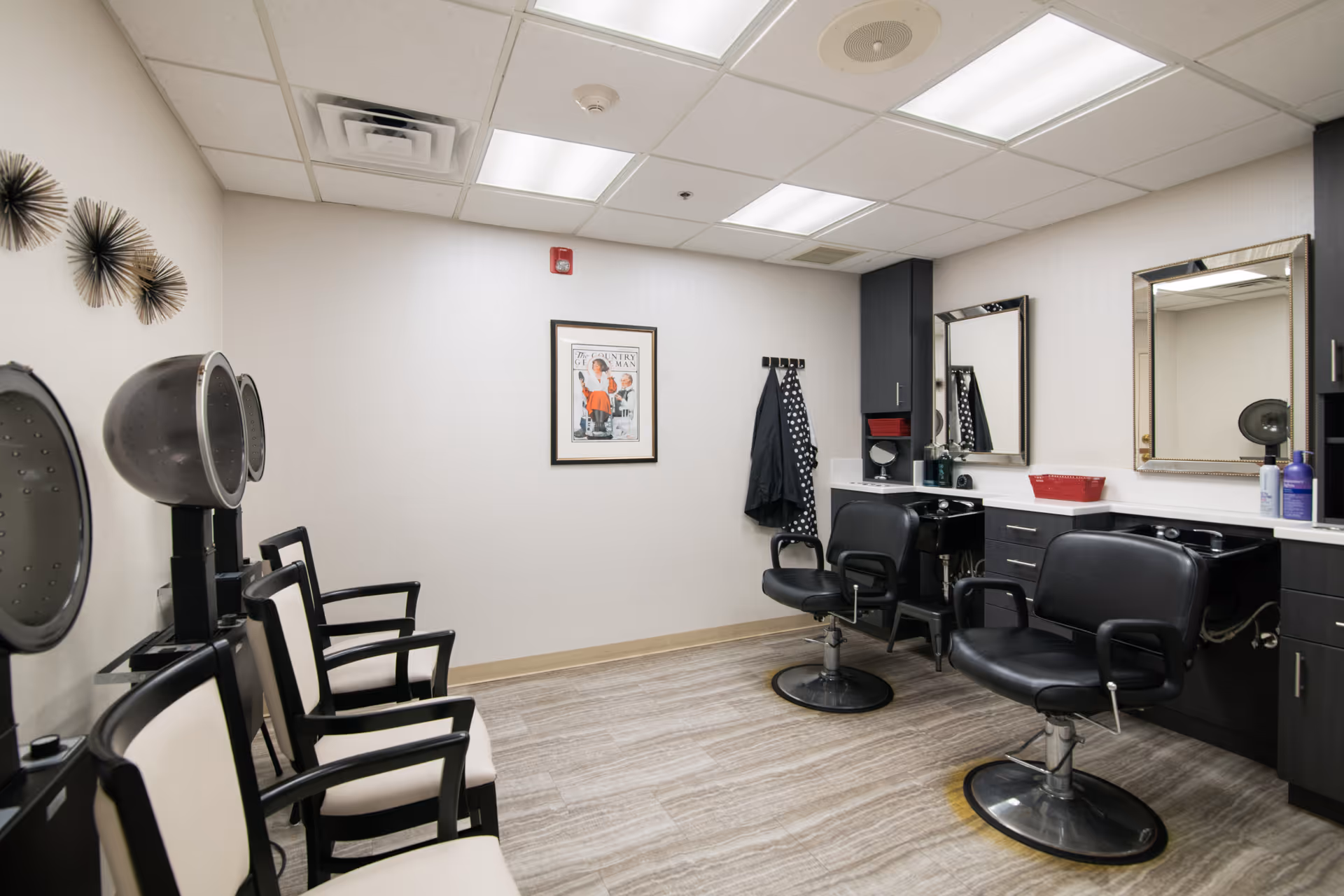 Interior of a hair salon room with black and white styling chairs, hair dryers, mirrors, and cabinets. The room has a light-colored floor and walls, with a framed picture on the wall and some hanging coats.