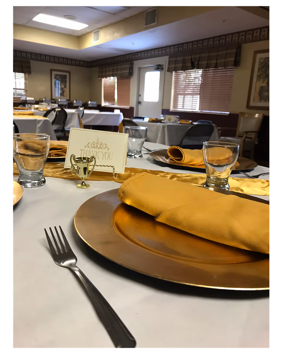 A dining room set up with tables covered in white tablecloths and gold runners. Each place setting includes a gold charger plate with a folded yellow napkin, a fork, and a glass. A small gold trophy holds a card that says 'Thank You'. The room has windows with blinds and framed artwork on the walls.