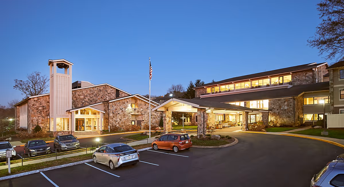 Exterior view of Ascension Living Alexian Village - Tennessee at dusk, showing a stone building with a covered entrance, an American flag on a flagpole, several parked cars, and illuminated windows.