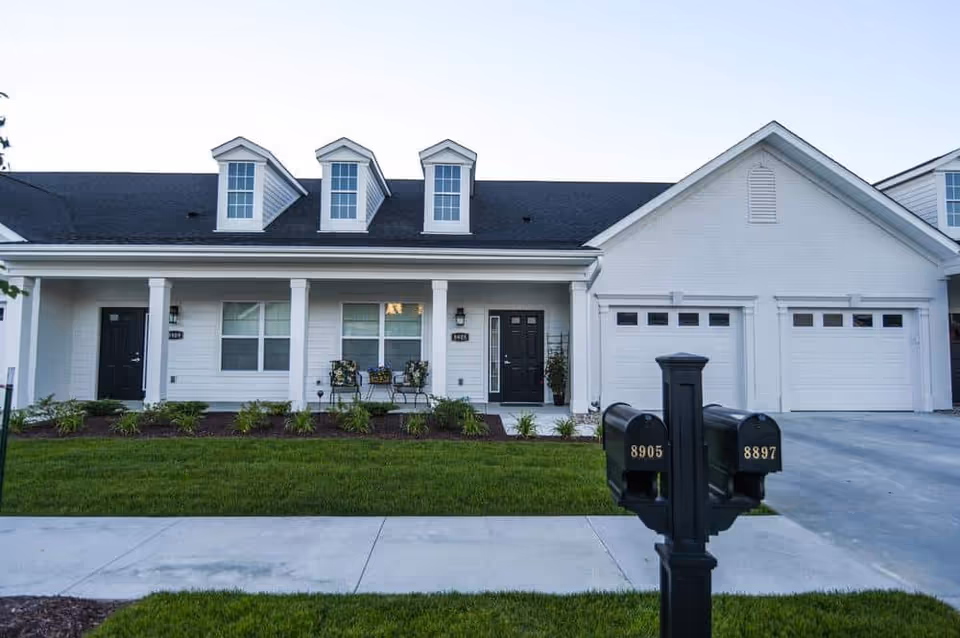 Exterior view of a single-story residential building with white siding, three dormer windows on the roof, a covered porch with two chairs and a small table, two black front doors, and two white garage doors. In the foreground, there are two black mailboxes with the numbers 8905 and 8897, a concrete sidewalk, and a well-maintained green lawn.