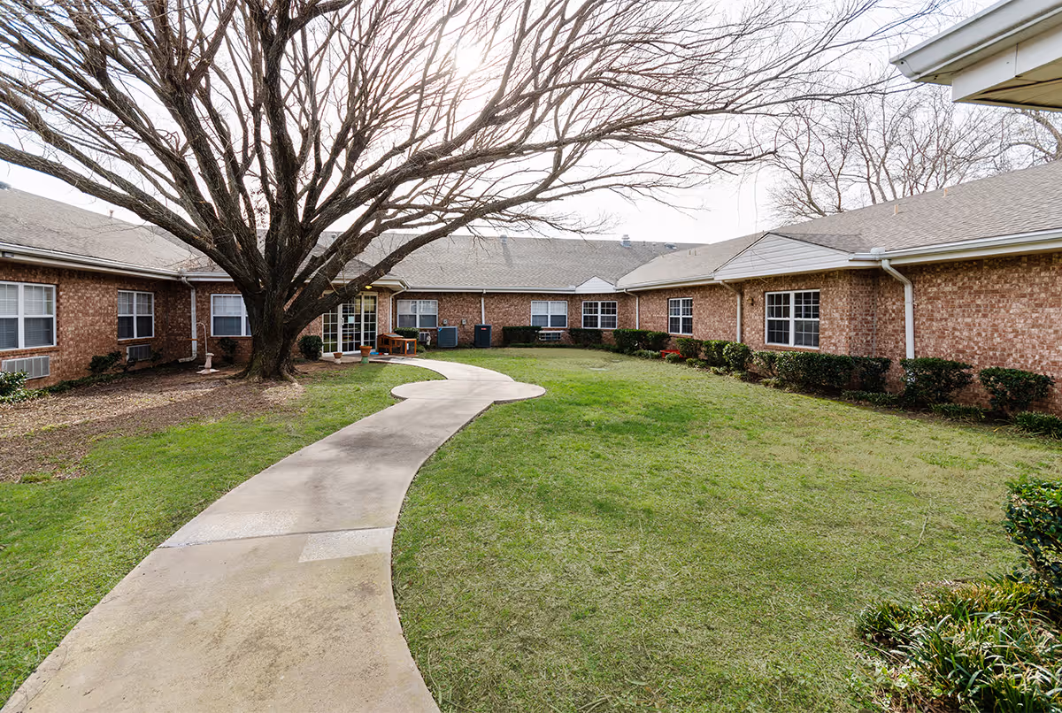 Curved concrete pathway leading through a grassy courtyard with a large leafless tree in the center, surrounded by a single-story brick building with multiple windows and a gray roof under an overcast sky.