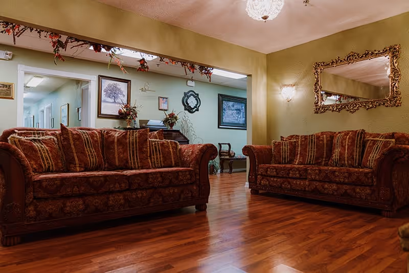 A cozy living room area in an assisted living facility featuring two ornate patterned sofas with multiple cushions, a large decorative mirror on the wall, framed artwork, and wooden flooring. The walls are painted green and there are floral decorations above the doorway.