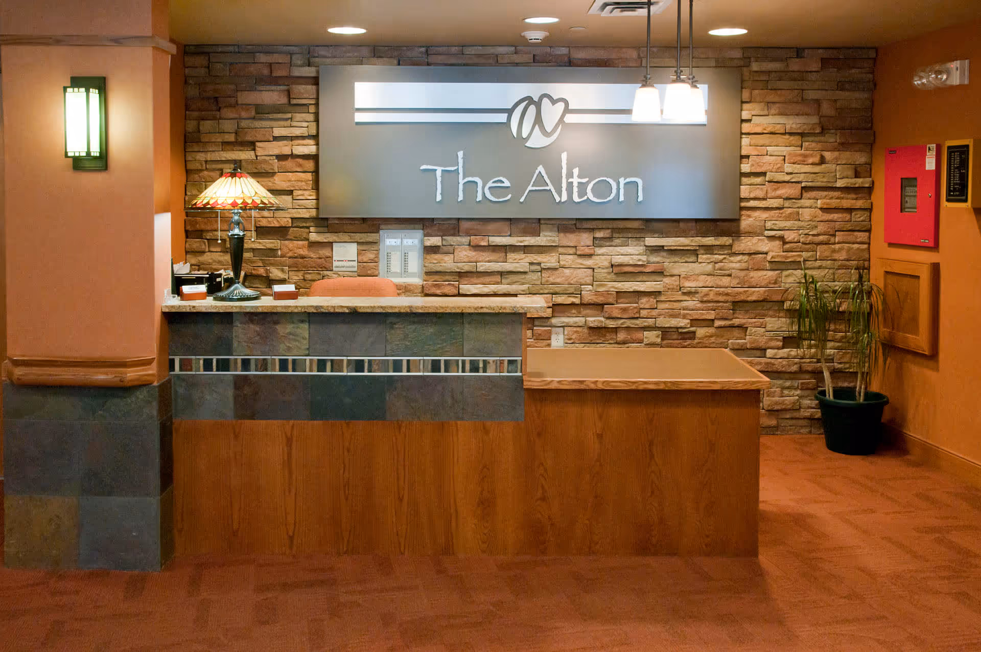 Reception desk area inside The Alton facility with a stone accent wall behind it, a sign displaying 'The Alton', a decorative lamp on the desk, a potted plant to the right, and warm lighting.