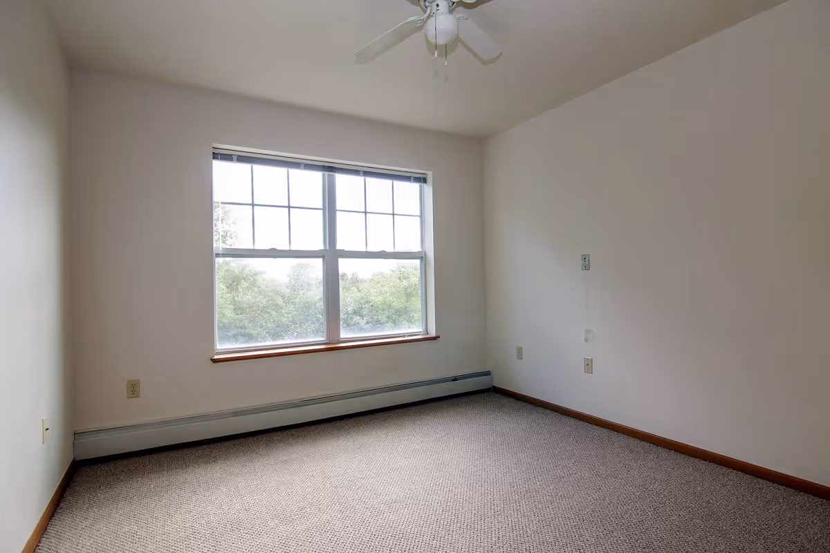 Empty room with beige carpet, white walls, a large window with a view of trees outside, and a white ceiling fan.