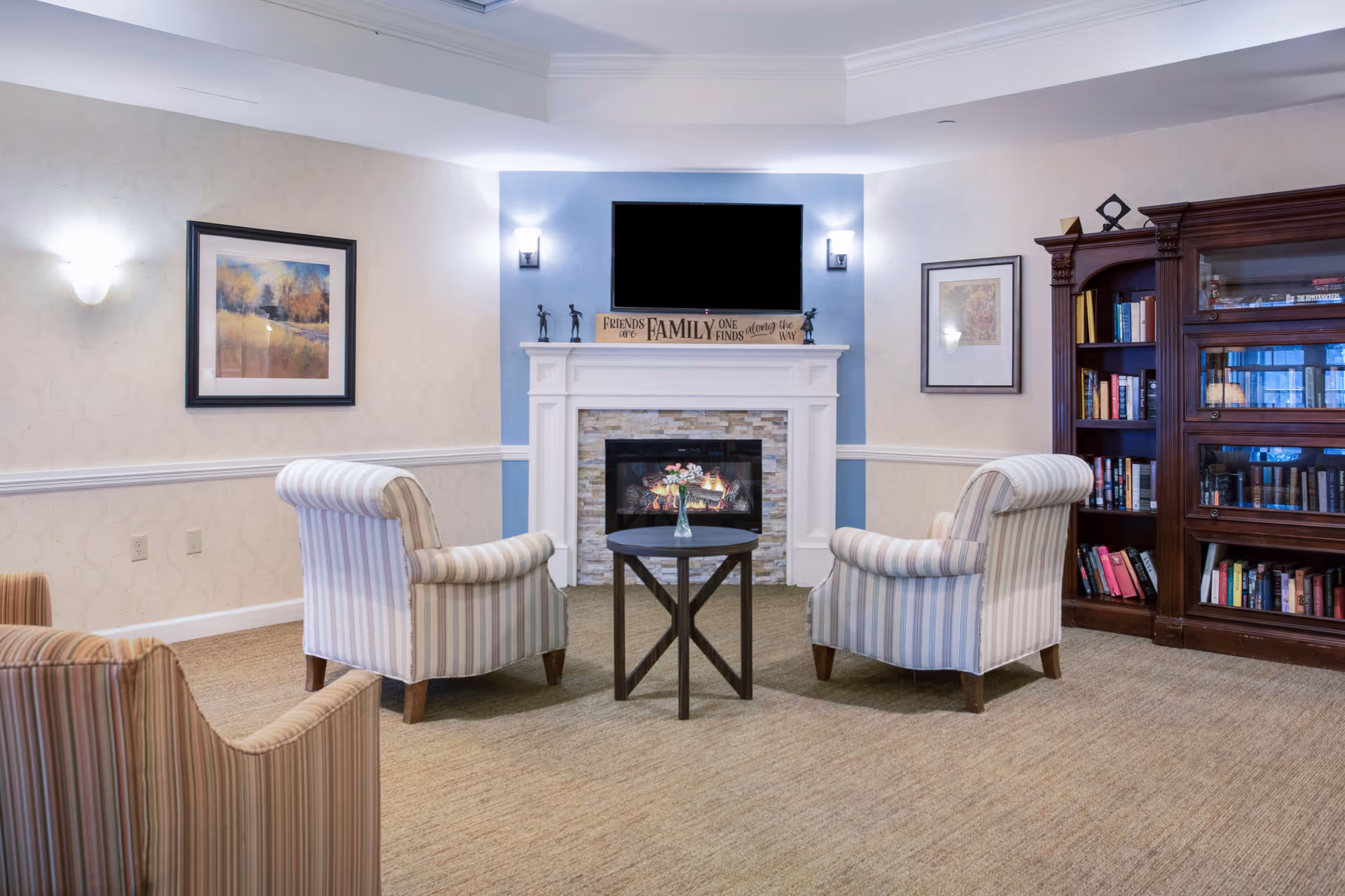 A cozy living room area with a fireplace in the center, above which is a mounted flat-screen TV. Two striped armchairs face the fireplace with a small round table between them holding a vase with flowers. On the right side, there is a wooden bookshelf filled with books. The walls are decorated with framed artwork and wall sconces providing soft lighting.