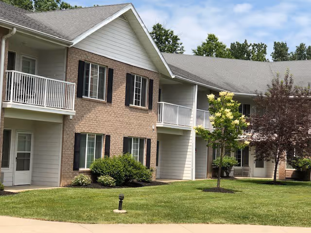 Exterior view of a two-story senior living facility building with brick and light-colored siding, balconies with white railings, several windows with black shutters, green grass lawn, and small trees under a partly cloudy sky.