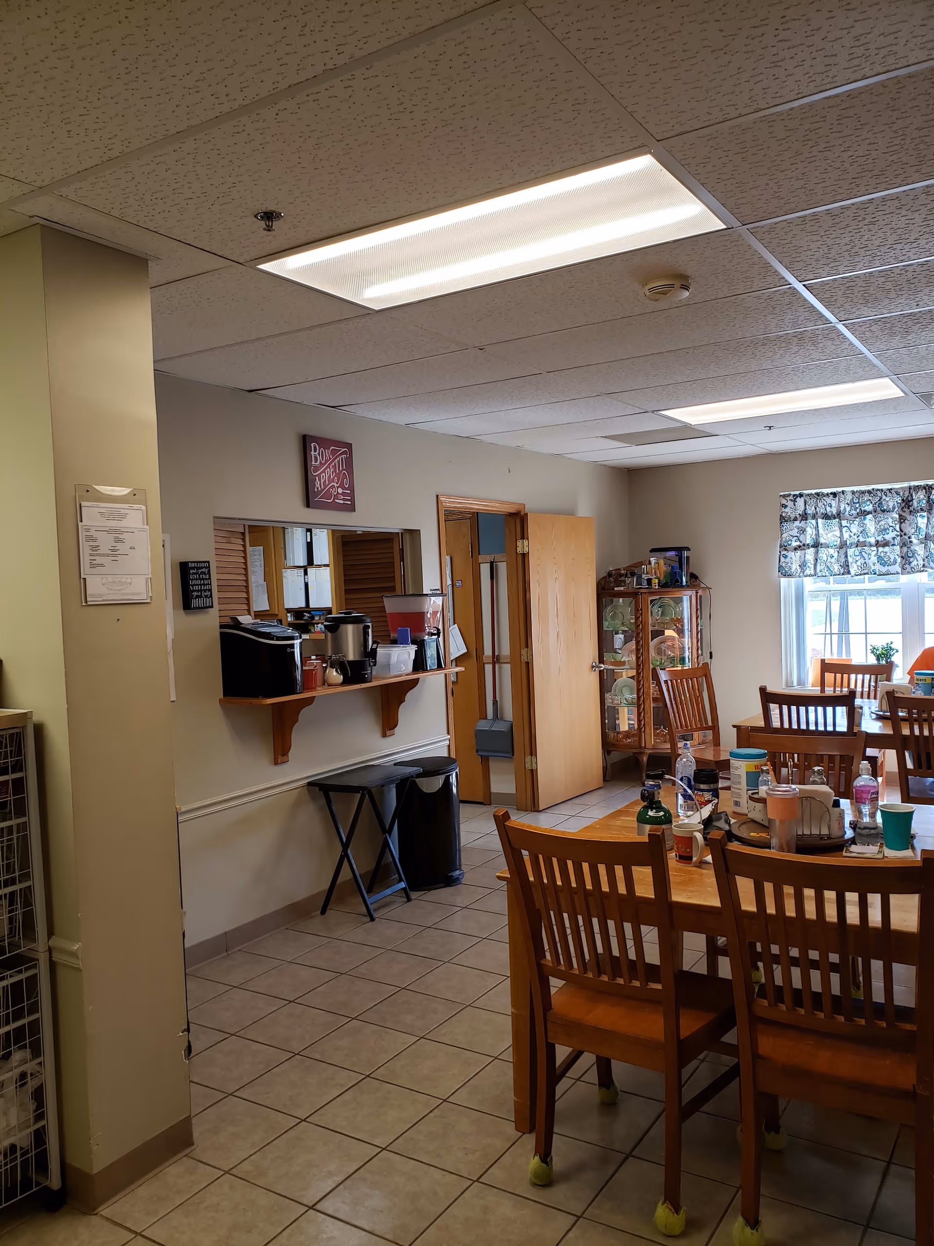 Interior view of a dining area in a senior living facility with wooden tables and chairs. The tables have various items on them including cups, bottles, and containers. There is a window with patterned curtains letting in natural light. On the left wall, there is a serving window with a shelf holding beverage dispensers and other items. The floor is tiled, and the ceiling has fluorescent lighting panels.