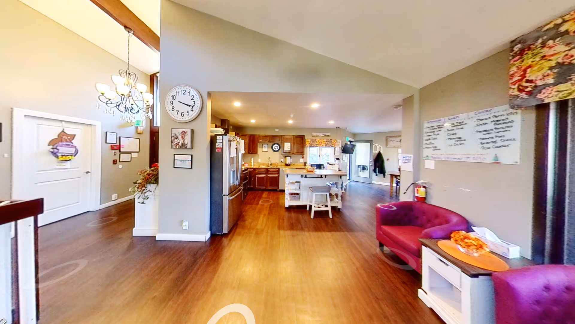 Open communal interior showing a kitchen island, refrigerator, seating areas, and a wall clock in a senior living facility.