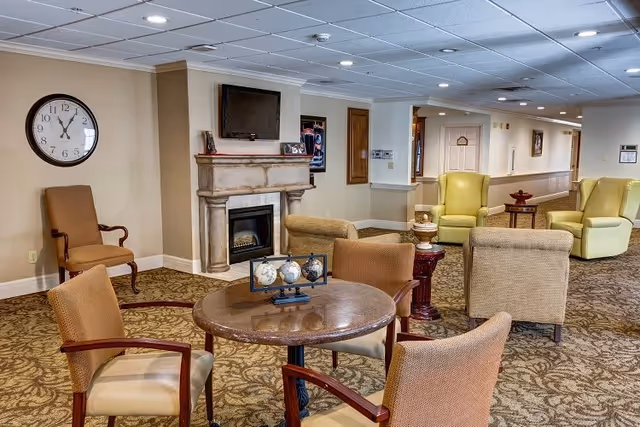 A cozy senior living facility common area with a round table and four chairs in the foreground. Behind the table is a fireplace with a mounted TV above it, flanked by framed pictures. There are several comfortable armchairs and side tables arranged around the room. A large wall clock shows the time as 11:55. The room has beige walls, patterned carpet, and a drop ceiling with recessed lighting.