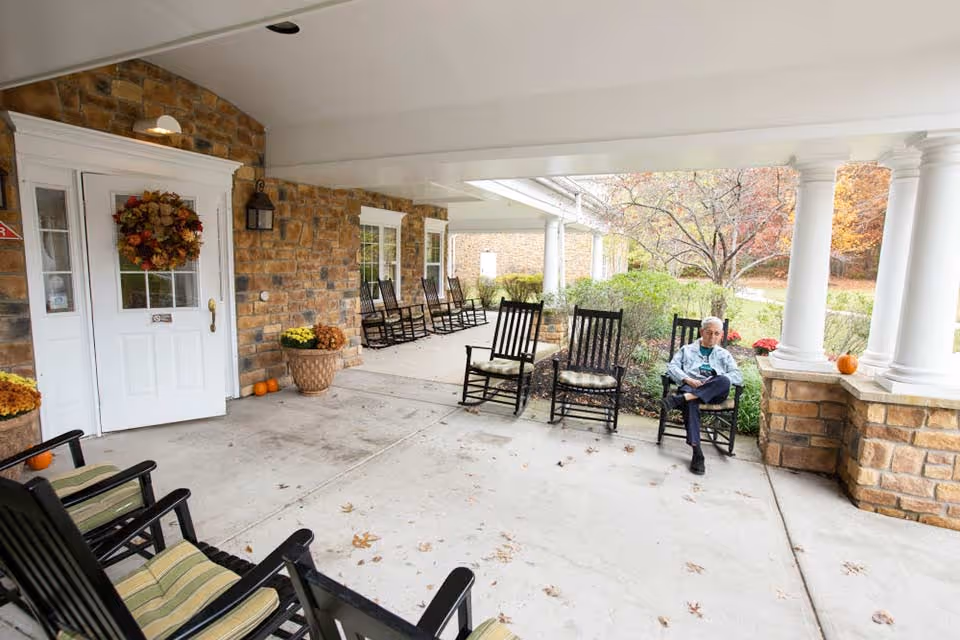 Covered stone entrance porch with multiple rocking chairs, autumn decorations, and an elderly man seated near white columns.