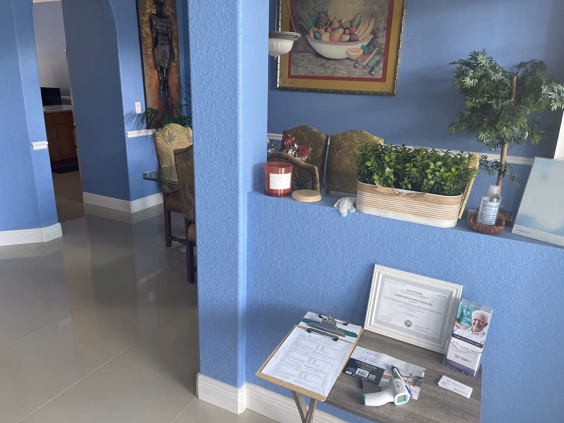Interior view of a senior living facility with blue walls and glossy tiled floor. A small table holds a framed certificate, brochures, a thermometer, and paperwork. There are decorative plants, a candle, and a painting of fruit on the wall. A dining area with upholstered chairs and a glass table is visible in the background.
