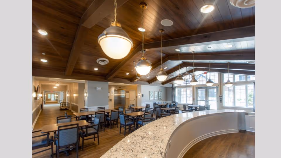 Spacious dining room with multiple wooden tables and chairs arranged neatly. The room features a high wooden ceiling with exposed beams and several hanging pendant lights. Large windows allow natural light to fill the space, and a curved granite countertop is visible in the foreground.