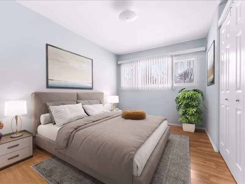 A neatly made bed with beige bedding and multiple pillows in a bright bedroom. There are two bedside tables with lamps on either side of the bed. A large window with vertical blinds lets in natural light. A green potted plant is placed near the window, and there is a framed artwork hanging above the bed. The floor is wooden with a gray rug under the bed.