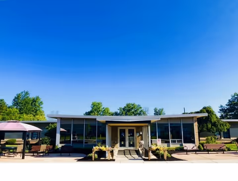 Front entrance of a single-story senior living building with benches, planters, and shaded patio seating beneath a clear blue sky.