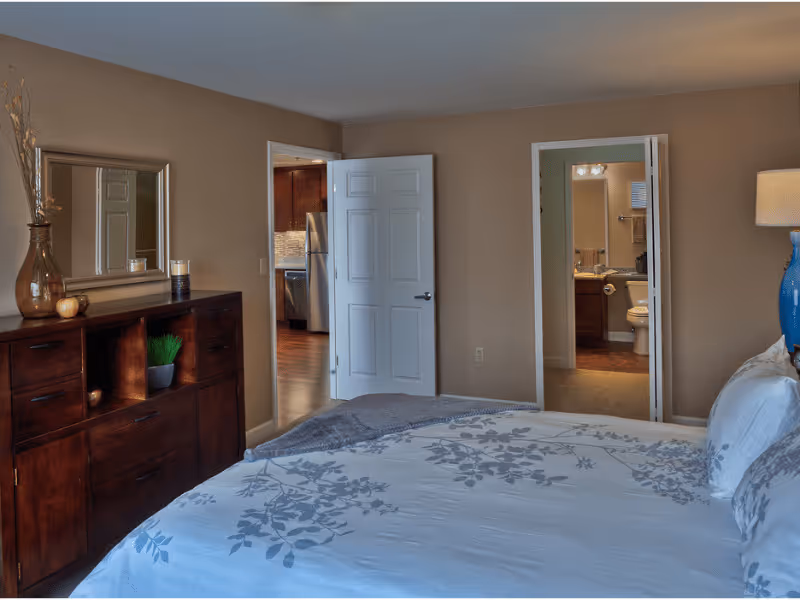 A bedroom with a bed covered in white and gray floral bedding, a wooden dresser with decorative items and a mirror above it, and two open doors showing a kitchen area with stainless steel appliances and a bathroom with a toilet and sink.