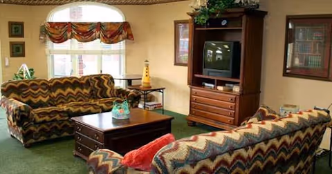 A cozy living room with two patterned sofas facing a wooden coffee table. There is a wooden entertainment center with a TV and drawers, a window with a valance, and framed pictures on the walls. The room has green carpeting and a warm, inviting atmosphere.