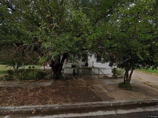 Front of a small white house partially hidden by large trees with a sidewalk, steps and a chain-link fence in front.