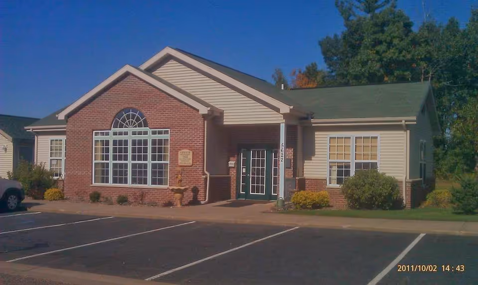Exterior view of a single-story brick and siding building with a large window and a green door entrance, surrounded by a parking lot and some greenery under a clear blue sky.