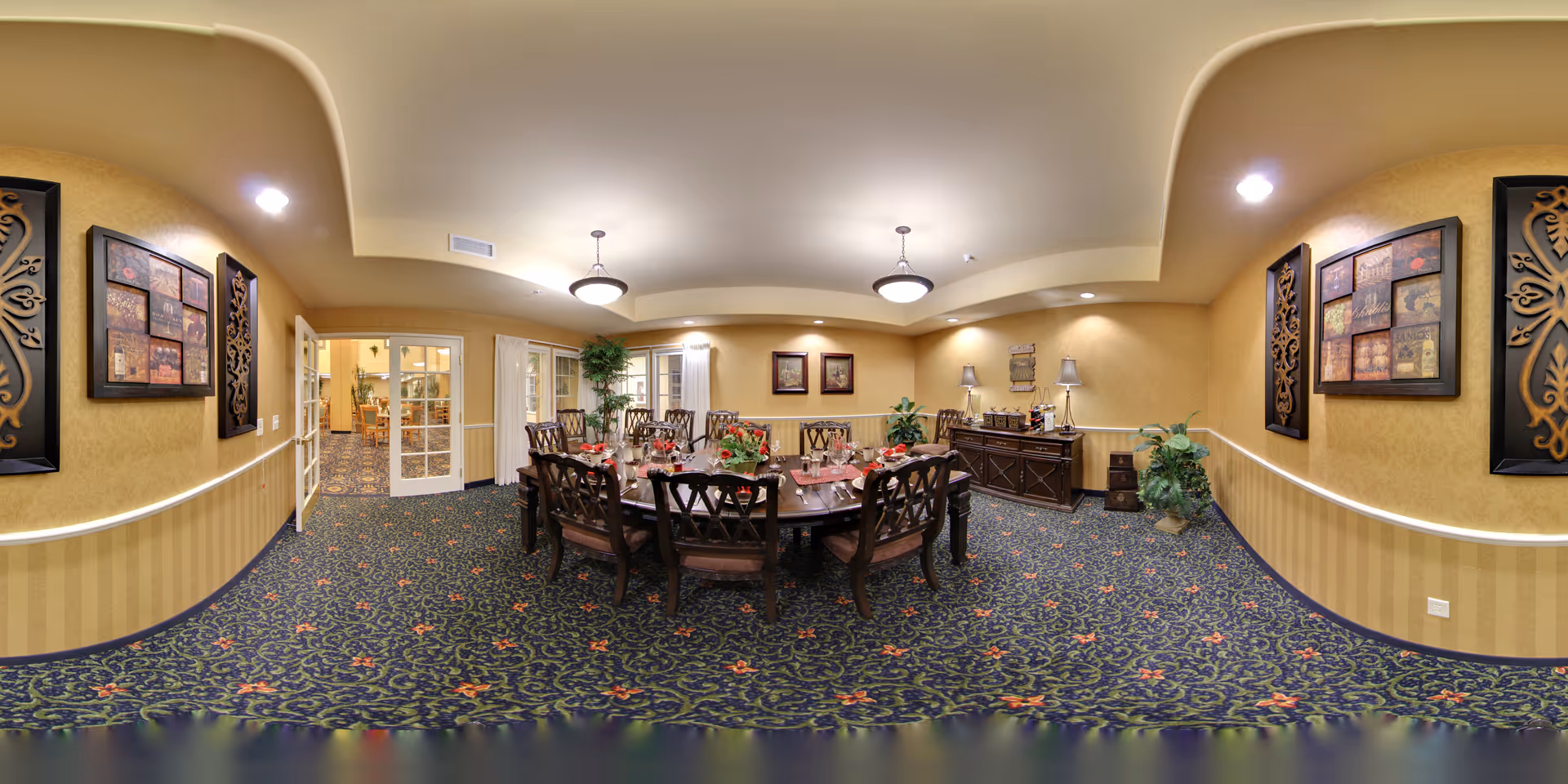A formal dining room with a large rectangular wooden table surrounded by ten chairs. The table is set with red napkins, plates, and floral centerpieces. The room has patterned carpet with floral designs, beige walls with framed artwork, and two ceiling light fixtures. There is a sideboard with lamps and decorative items against the far wall, and a potted plant in the corner. French doors lead to another room.