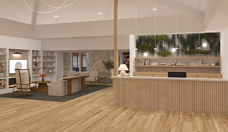A spacious, modern interior common area with light wood flooring and a wooden reception or service counter on the right. Behind the counter is a wall with hanging plants and pendant lights. To the left, there is a seating area with armchairs, a small table with a flower vase, built-in shelves, and a stone accent wall in the background.