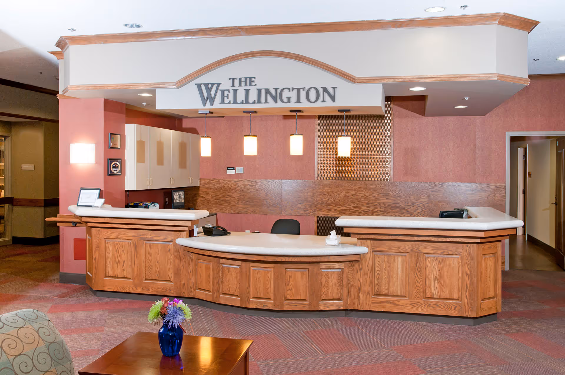 Curved wooden reception desk in a lobby under a sign reading "The Wellington," with pendant lights and a small table with a vase of flowers in the foreground.