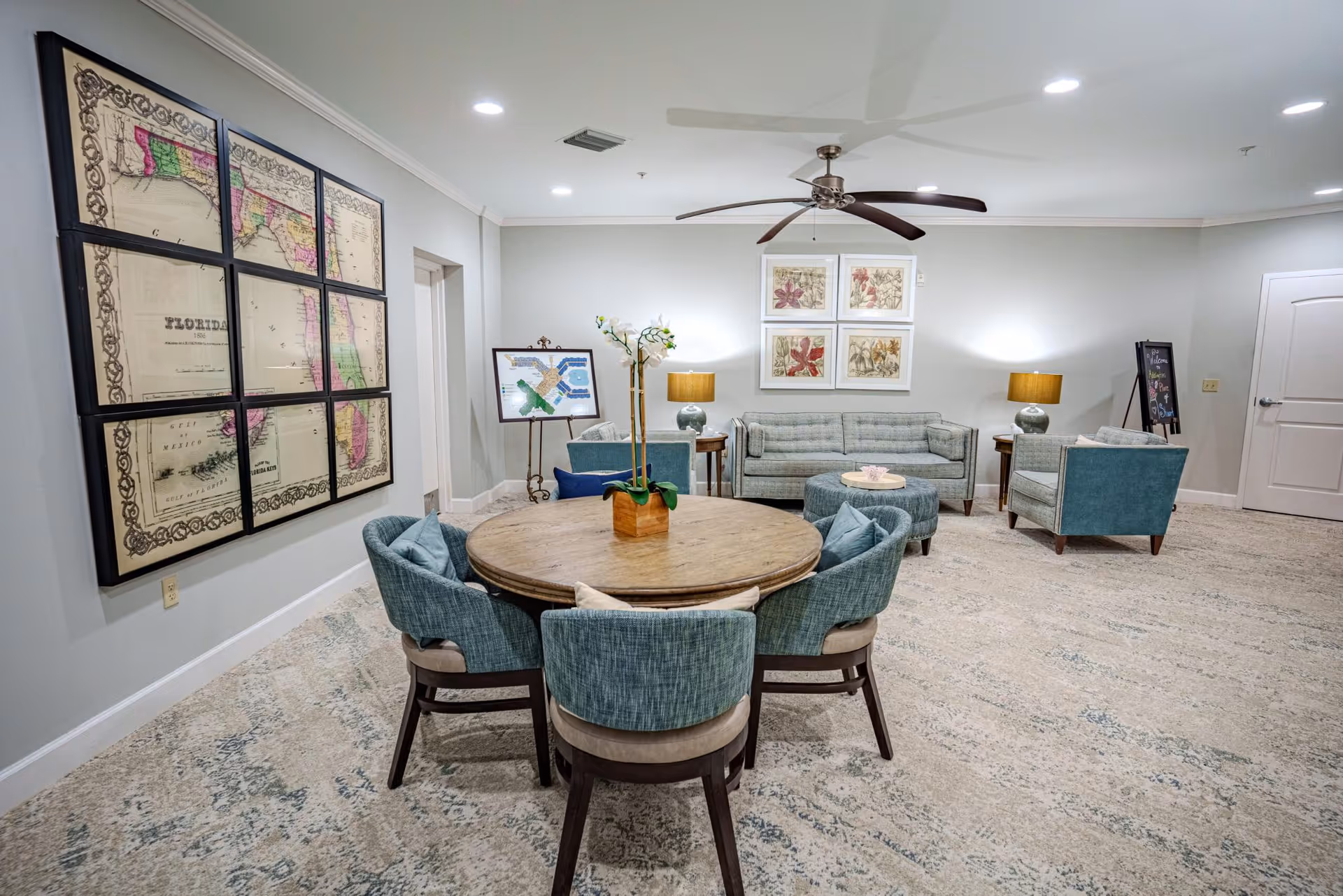 A cozy common area in a senior living facility featuring a round wooden table surrounded by six blue upholstered chairs. In the background, there is a seating area with a light blue sofa, two matching armchairs, and a round ottoman. The walls are decorated with framed floral artwork and a large vintage map of Florida. The room is well-lit with ceiling lights and two table lamps, and a ceiling fan is mounted above.