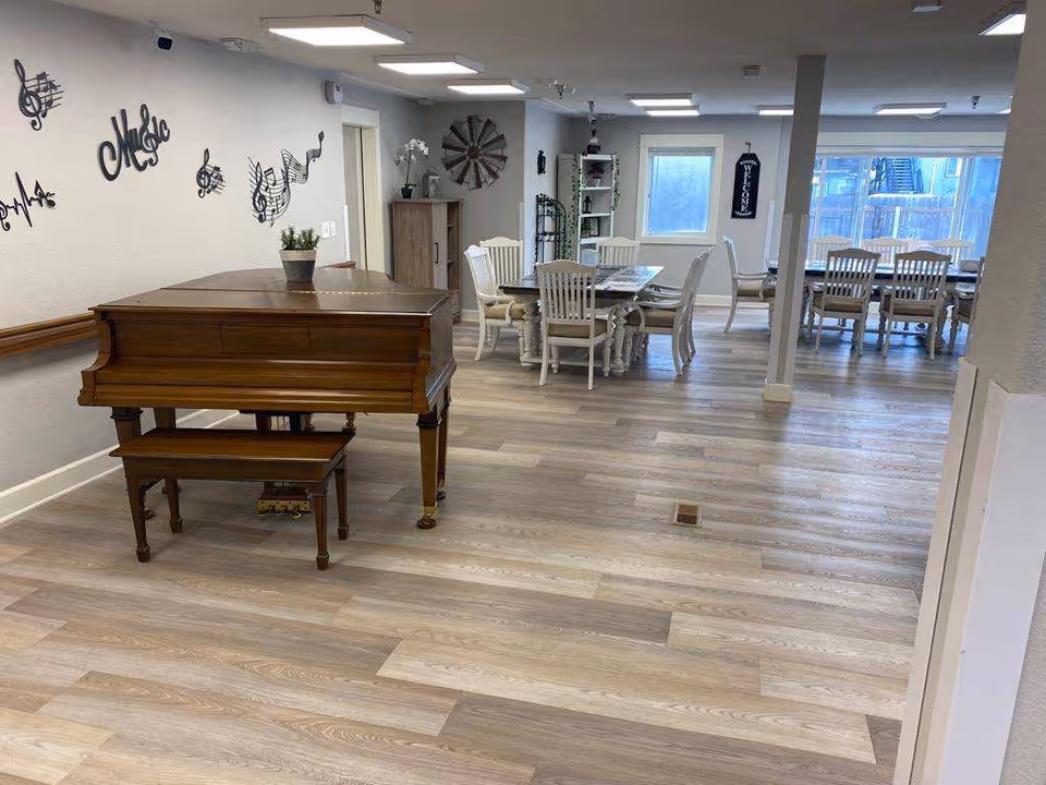 Interior of a memory care facility room with a wooden piano and bench on the left side, musical note wall decorations above the piano, and two dining tables with chairs in the background near windows. The room has light wood flooring and neutral-colored walls with ceiling lights.