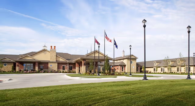Front exterior of a single-story senior living facility with a lawn, flagpoles, lamp posts, and a covered entrance.