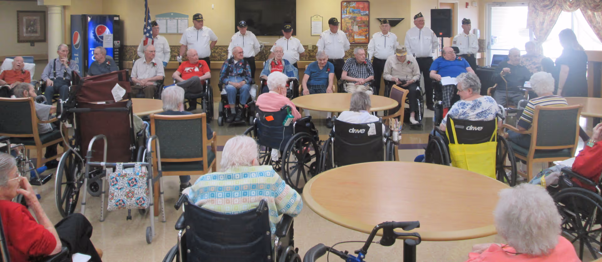 A group of elderly people, many in wheelchairs, seated in a common room facing a line of veterans standing in front of an American flag and a Pepsi vending machine. The room has round tables and chairs, with a woman standing on the right side.