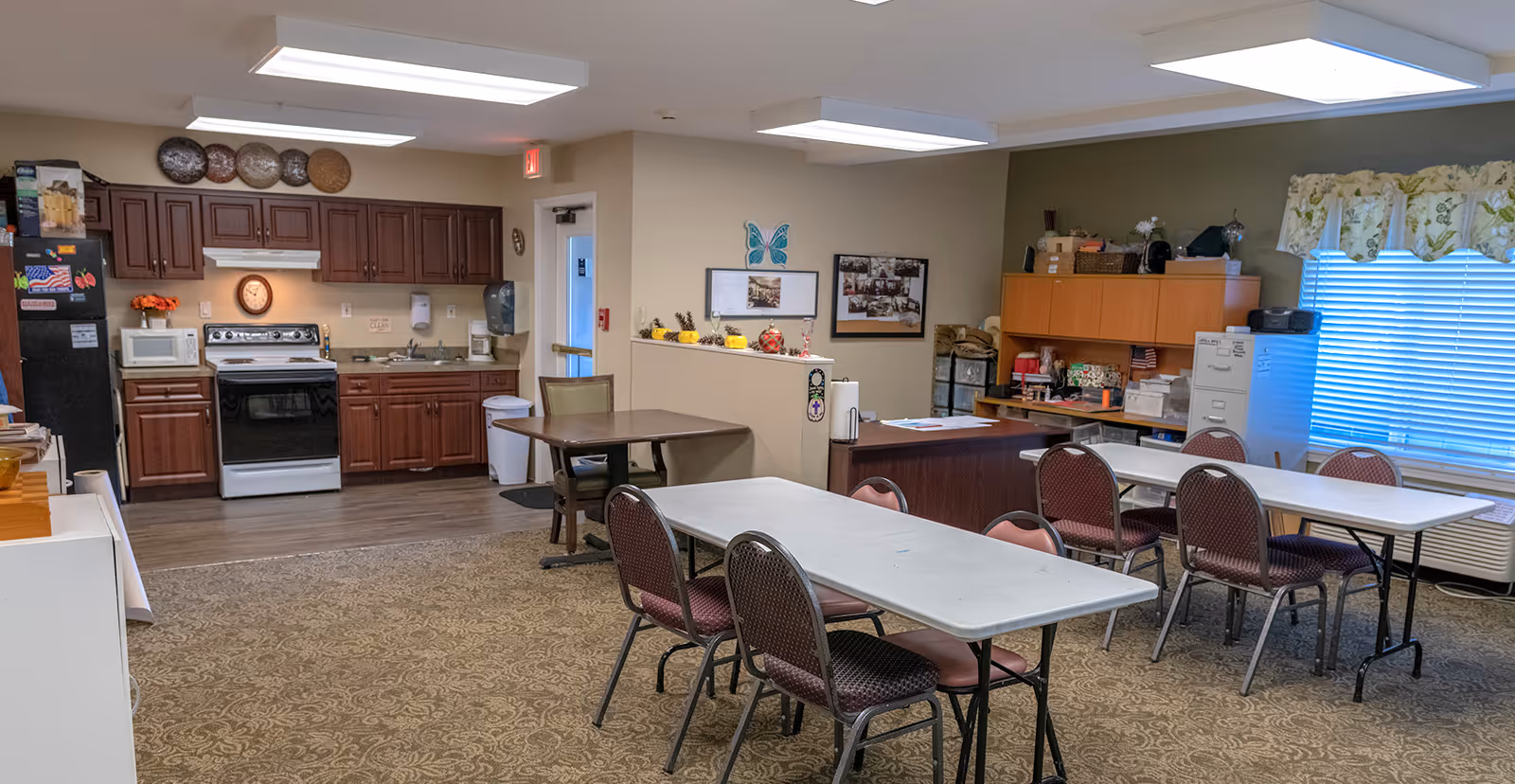 A communal room in a senior living facility featuring a kitchen area with dark wood cabinets, a stove, microwave, and refrigerator. In front of the kitchen are two folding tables with several chairs arranged around them. The room has carpeted flooring, fluorescent ceiling lights, and a window with floral curtains. There are various decorations and storage cabinets along the walls.