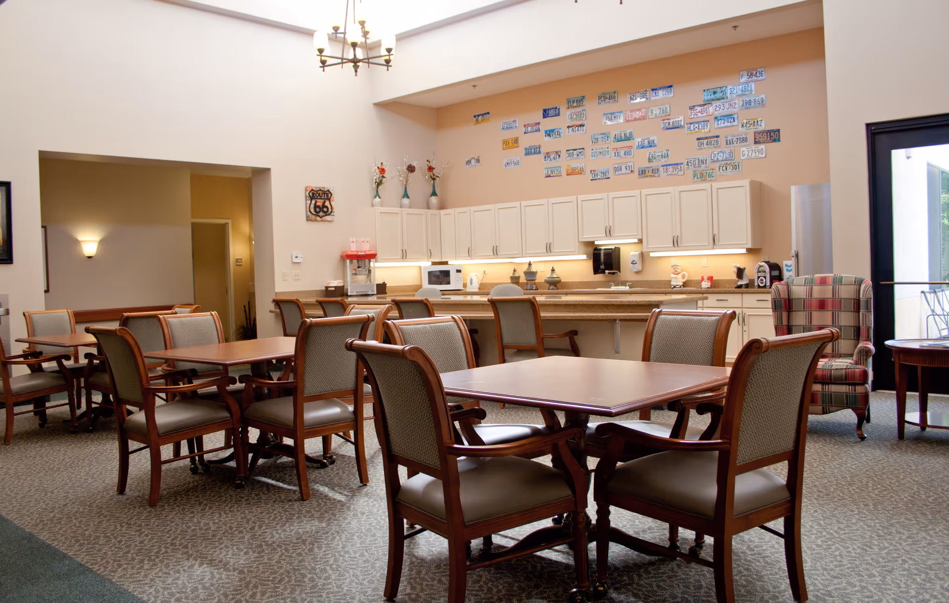 A bright communal dining area with several wooden tables and cushioned chairs. The back wall features a kitchenette with white cabinets, a microwave, a popcorn machine, and various decorative items. Above the cabinets, there are numerous colorful license plates displayed on the wall. A large window or glass door is visible on the right side, allowing natural light to enter the room.