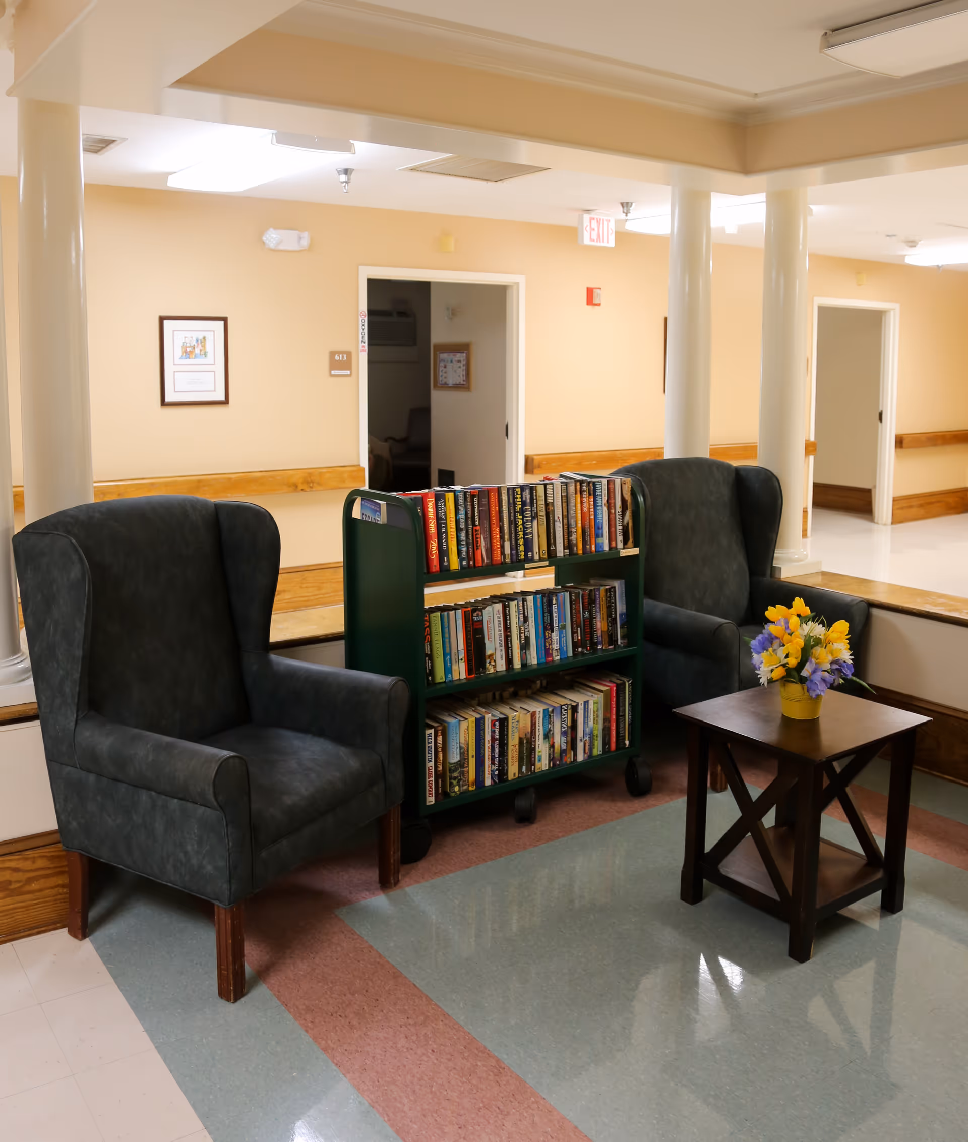 Two wingback chairs with a rolling book cart between them and a small table with a vase of flowers in a senior living facility hallway seating area.
