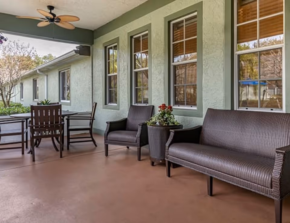Covered outdoor patio area with brown wicker seating including a loveseat and two armchairs, a small table with a flower pot, and a dining table with chairs. The patio has a ceiling fan and is adjacent to a light green building with several windows.