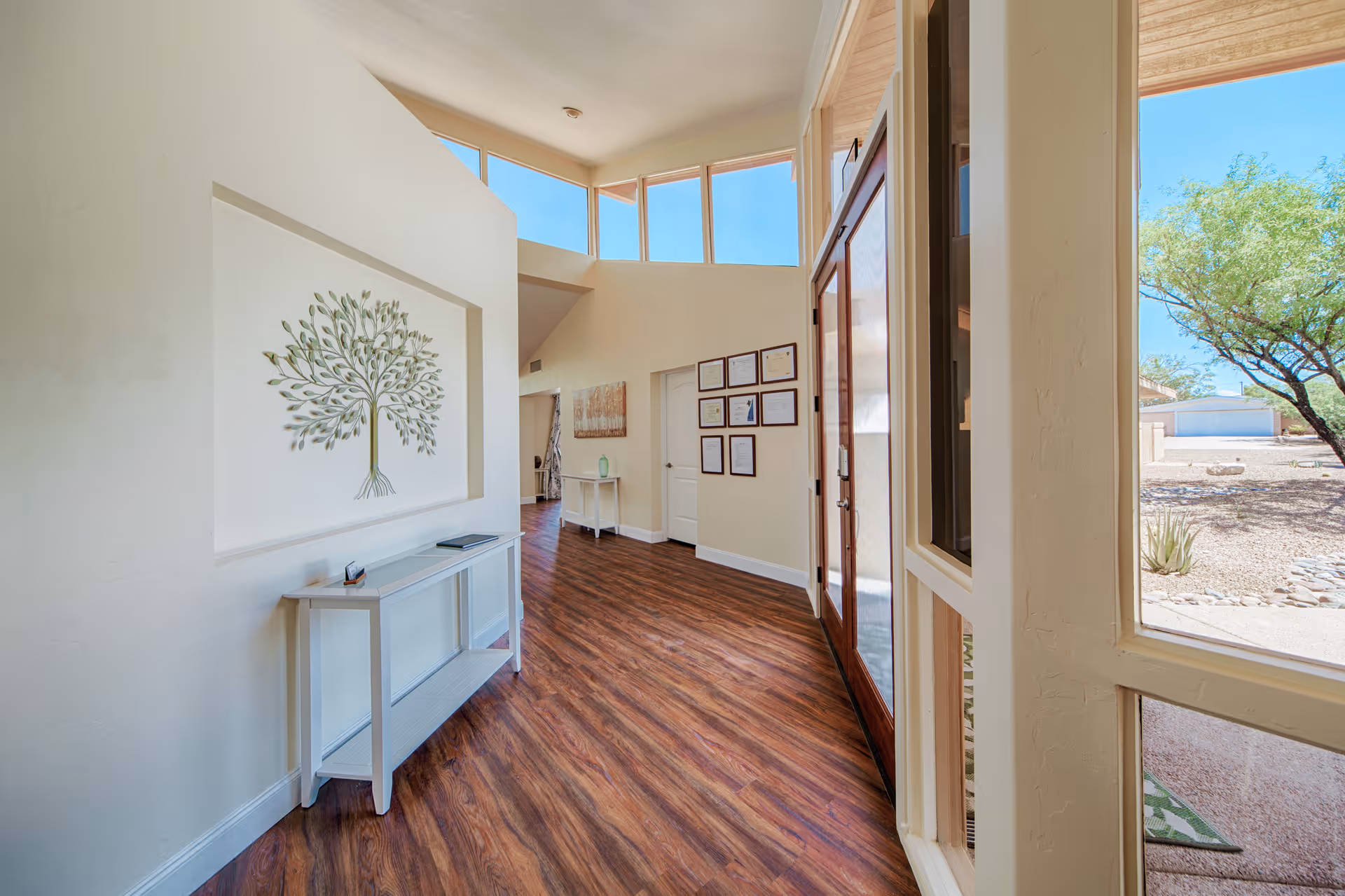 Bright hallway with wood flooring and cream-colored walls featuring a decorative tree wall art. There is a small white console table with a few items on it along the left wall. On the right side, large windows and a glass door provide a view of an outdoor area with desert landscaping and a tree. Several framed certificates or artworks are hung on the far wall.