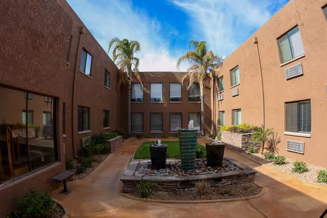 Sunlit enclosed courtyard of a two-story brown stucco facility with a central fountain, palm trees, walkways, and surrounding windows.