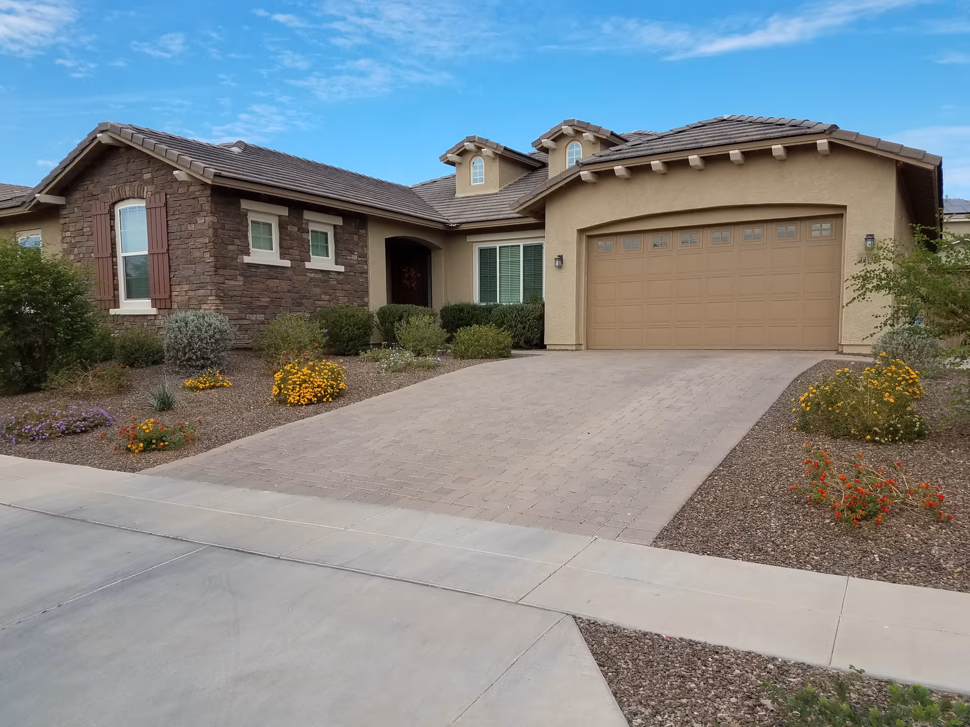 Exterior view of a single-story house with a stone and stucco facade, a two-car garage with a closed door, a paved driveway, and landscaped front yard with various shrubs and flowering plants under a blue sky.