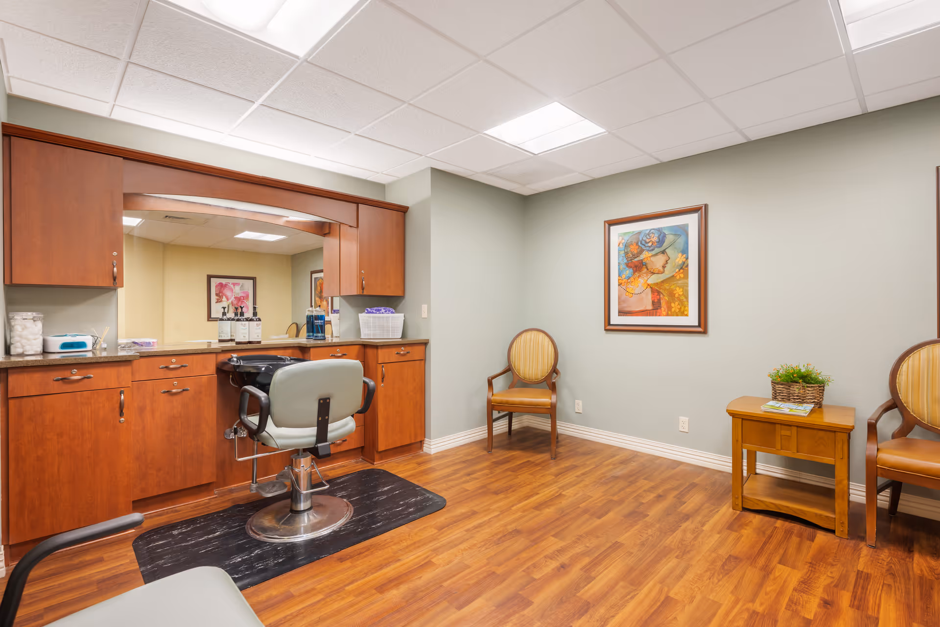 Interior view of a salon area in a senior living facility with a salon chair in front of a large mirror and wooden cabinetry. The room has wooden flooring, two upholstered chairs, a small wooden table with a plant, and a colorful framed painting on the wall.