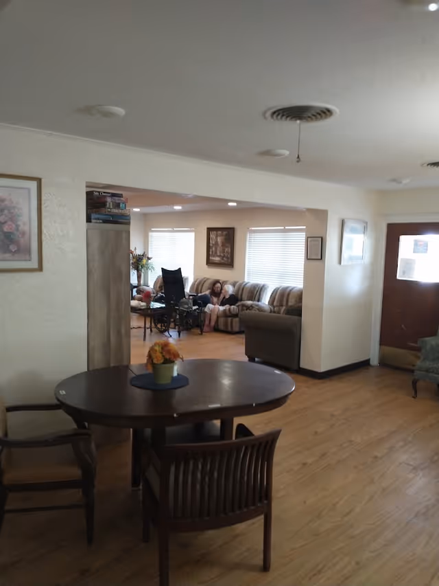 Interior view of a senior living facility common area with wooden flooring, a round wooden table with chairs, a flower pot centerpiece, and a seating area with couches and a wheelchair. A person is sitting on one of the couches near the windows with blinds.