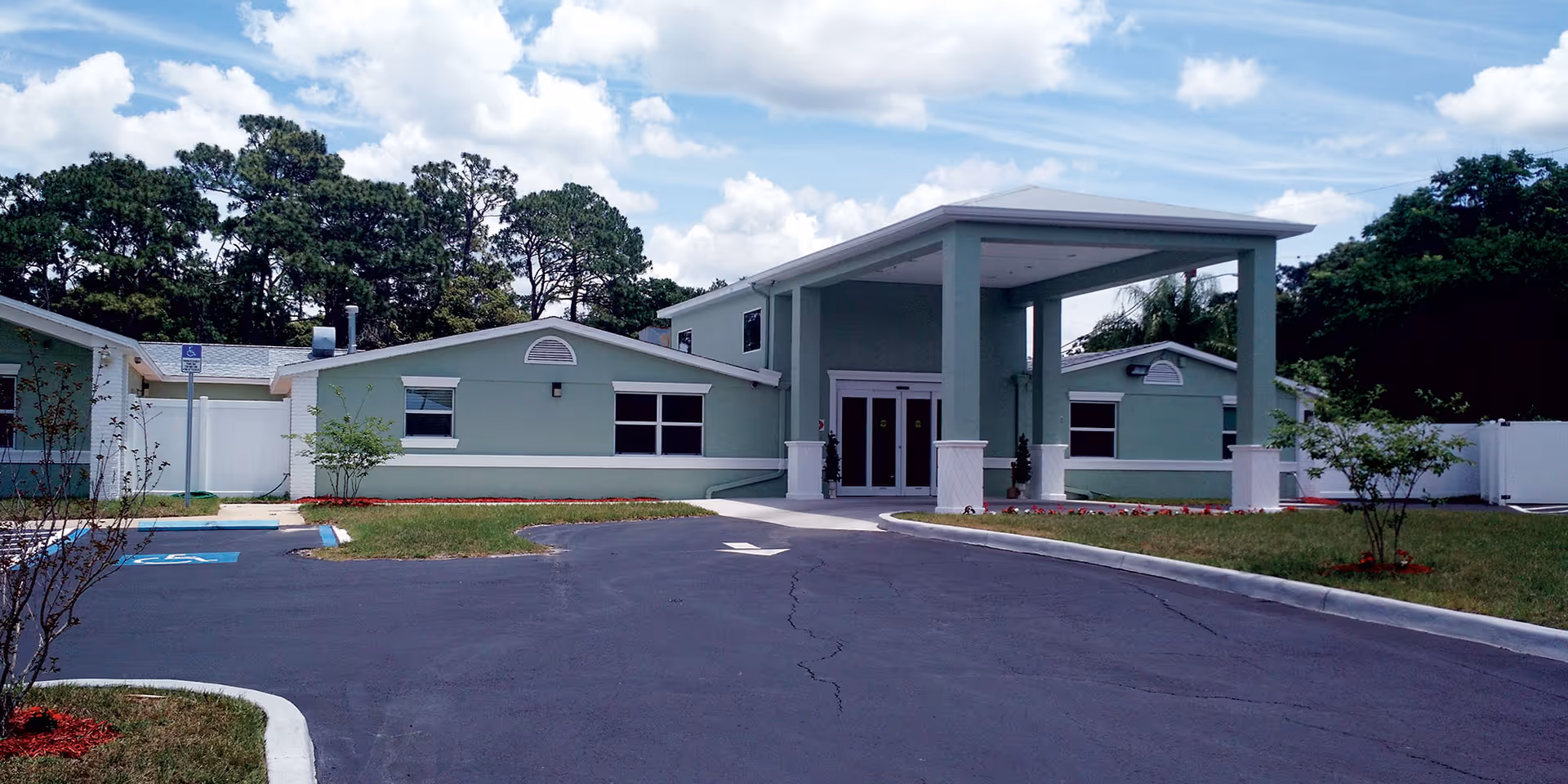 Front entrance of a low-rise light-green senior living facility with a covered porte-cochère, parking lot, and landscaping under a partly cloudy sky.