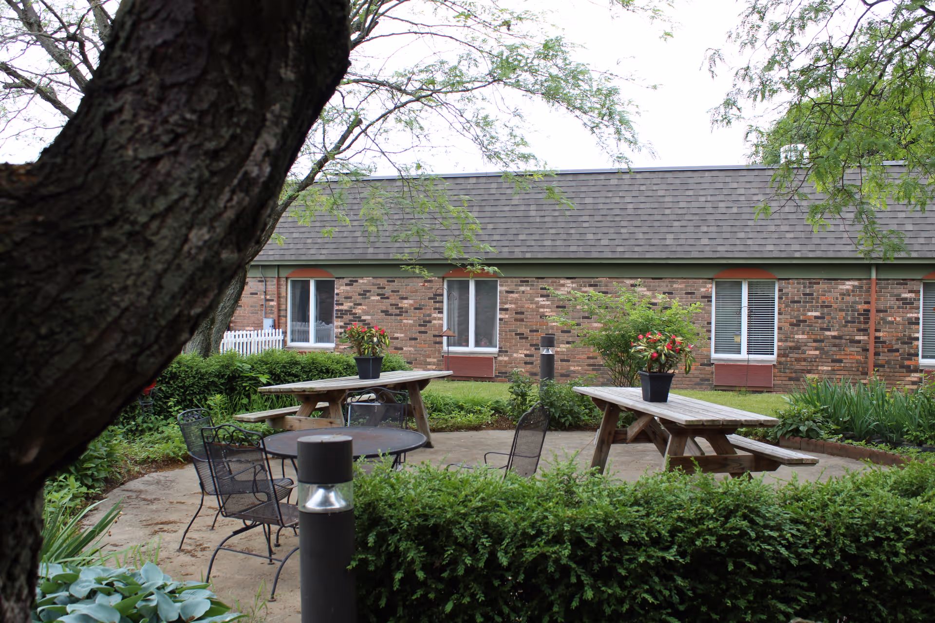 Outdoor courtyard with picnic tables, metal chairs, potted flowers, and shrubbery in front of a single-story brick building.