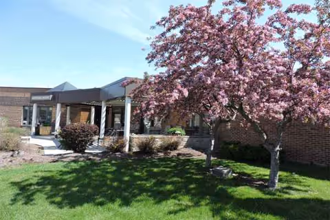 Brick senior living facility entrance with a covered walkway, pink flowering trees, and green lawn