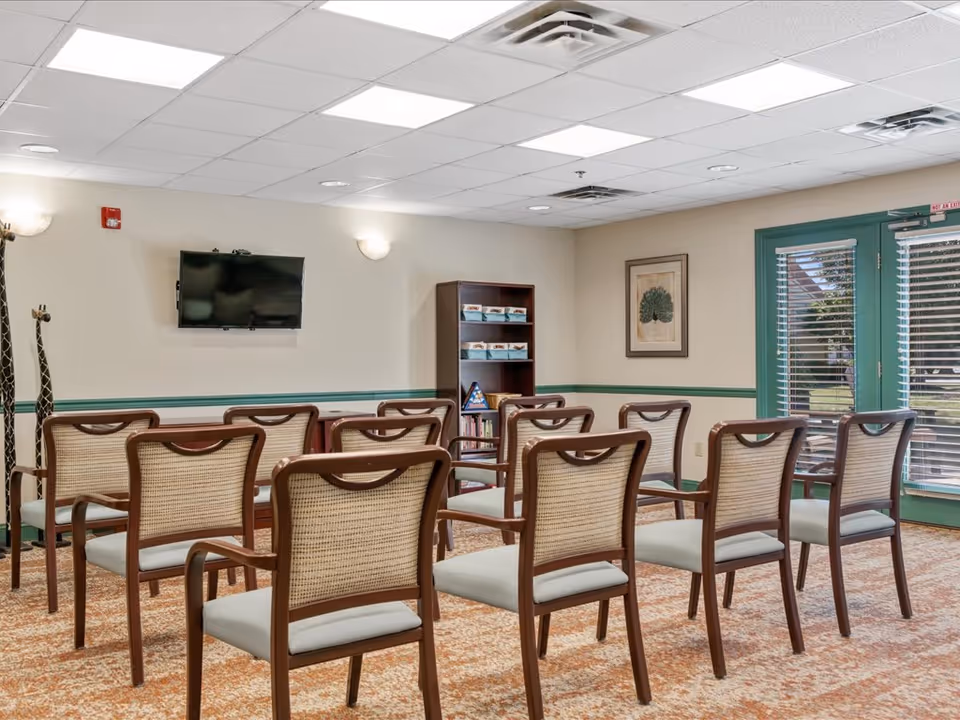 A small meeting or activity room with two rows of wooden chairs with woven backs facing a wall-mounted flat screen TV. The room has beige walls with green trim, a carpeted floor, a bookshelf with books and games, a framed picture on the wall, and double glass doors with blinds leading outside.
