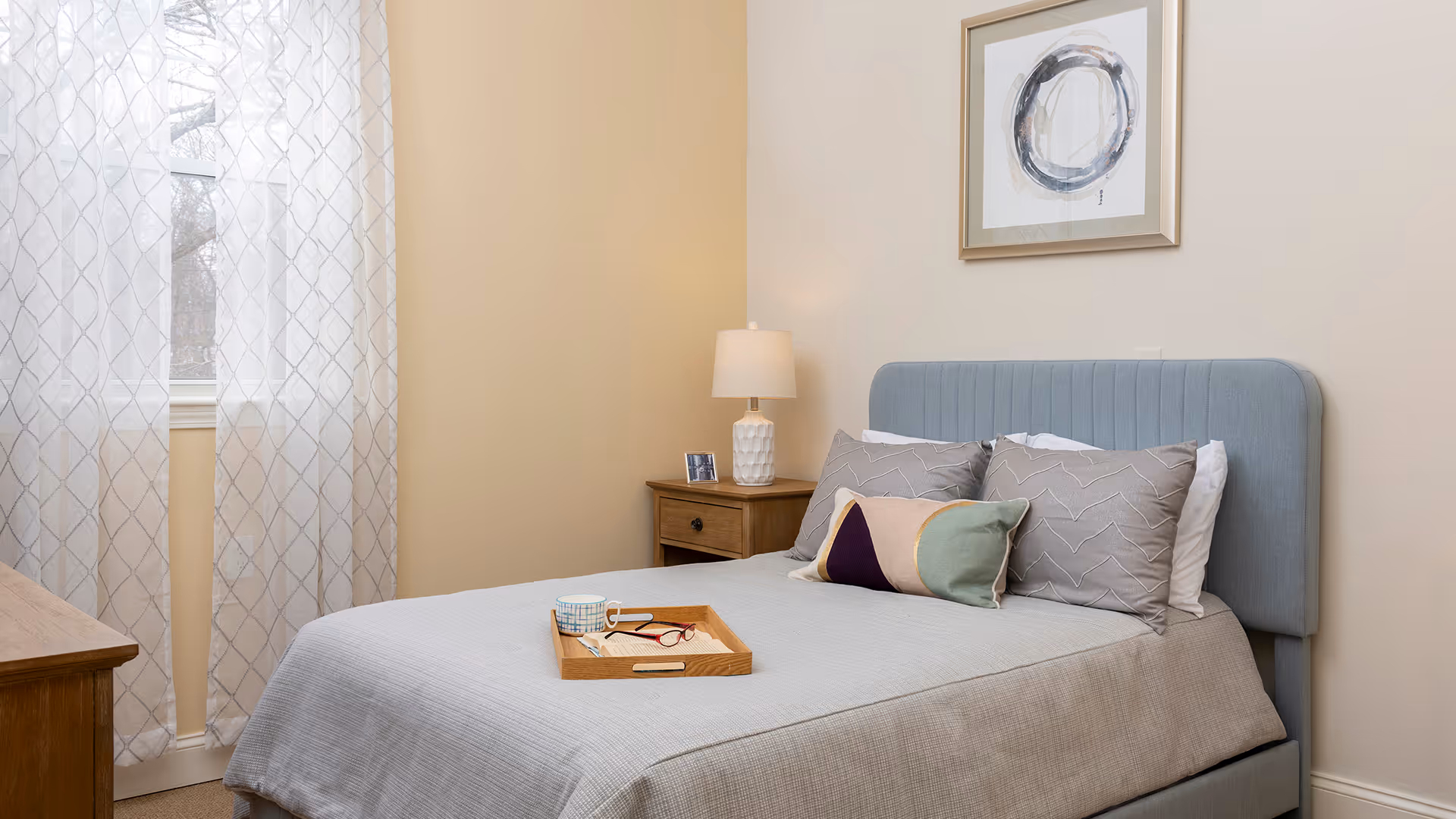 A neatly made bed with a light blue upholstered headboard in a bedroom. The bed has a gray bedspread, three pillows, and a decorative cushion. A wooden nightstand with a white lamp and a small framed photo is beside the bed. Sheer white curtains cover a window on the left side, and a framed abstract artwork hangs on the wall above the bed.