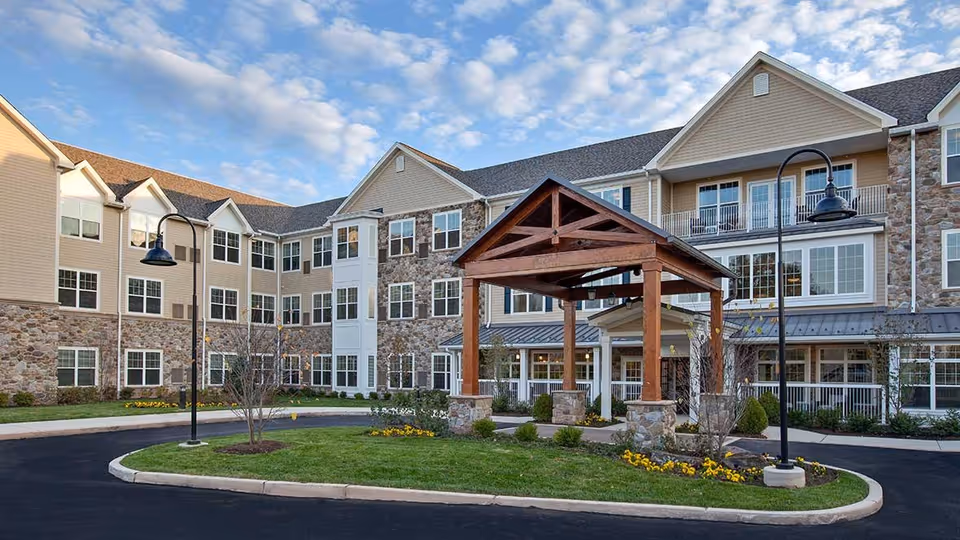 Exterior view of The Solana Doylestown senior living facility showing a multi-story building with stone and beige siding, numerous windows, a covered entrance with wooden beams, landscaped greenery, and a curved driveway under a partly cloudy sky.
