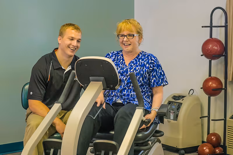 A woman in a blue patterned blouse is seated on a stationary exercise bike, smiling, while a man in a black polo shirt sits beside her, also smiling. They appear to be in a rehabilitation or healthcare facility room with exercise equipment and a rack holding red exercise balls in the background.
