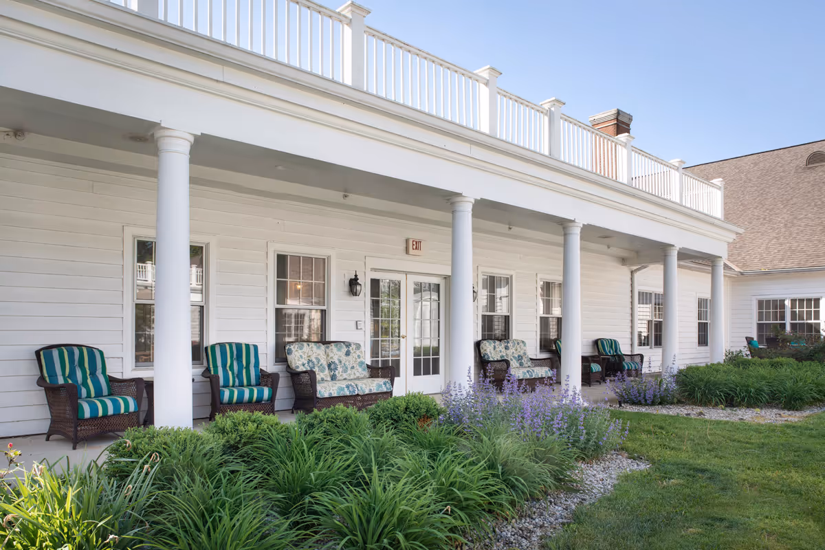Outdoor patio area of a senior living facility with white columns supporting an upper balcony. Several wicker chairs and loveseats with striped and floral cushions are arranged along the porch. There are green shrubs and purple flowering plants in the garden bed in front of the patio, and the sky is clear and blue.