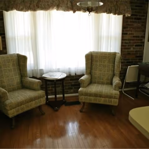 Two upholstered armchairs with a checkered pattern are placed on a wooden floor in front of a window with sheer white curtains and a floral valance. A small round wooden side table is positioned between the chairs. The room has a brick wall and a ceiling light fixture above.