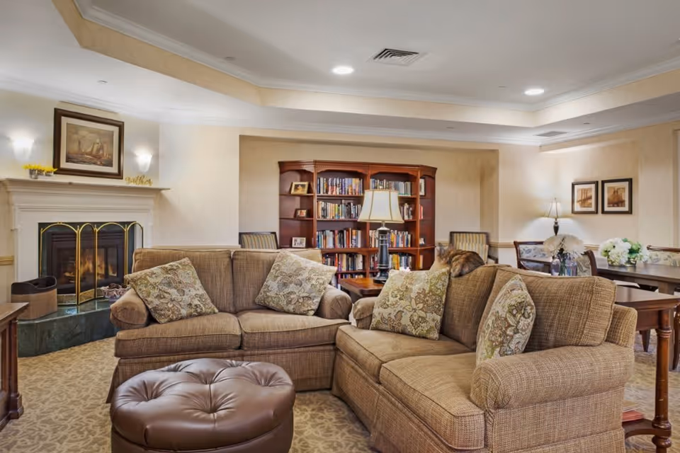 A cozy living room area with two beige sofas adorned with patterned cushions, a round brown leather ottoman, a wooden bookshelf filled with books, a table lamp, and a fireplace with a painting above it. The room also features a dining table with chairs and floral arrangements, soft lighting, and neutral-colored walls.